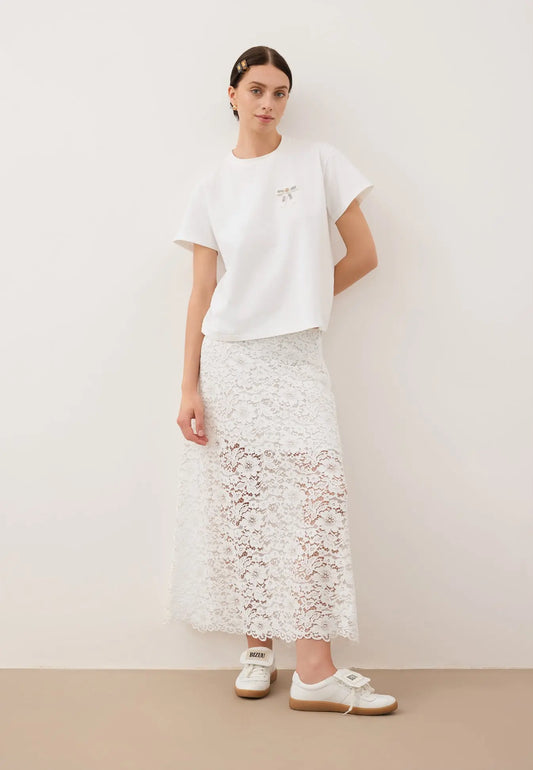 Modelka oparta o ścianę z jedną ręką zgiętą do tyłu ubrana w T-shirt z logowaną kokardką TISHALLE, białą, koronkową spódniczkę MILKY i buty MOBBY.