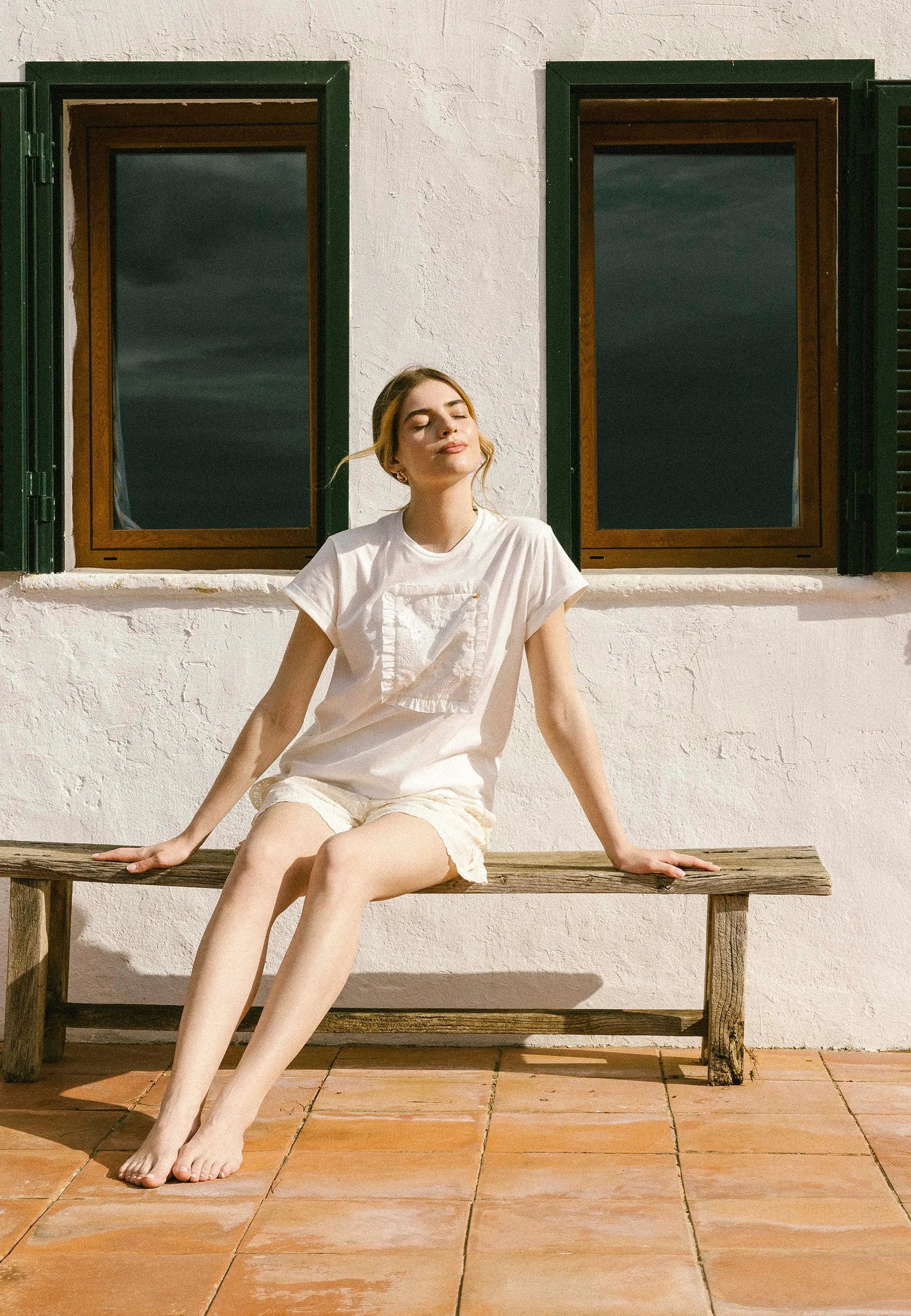 Modelka siedząca na drewnianej ławce na tle kolorowej ściany i okien w kremowym T-shircie MIRADO i szortach z koronki NILDA.