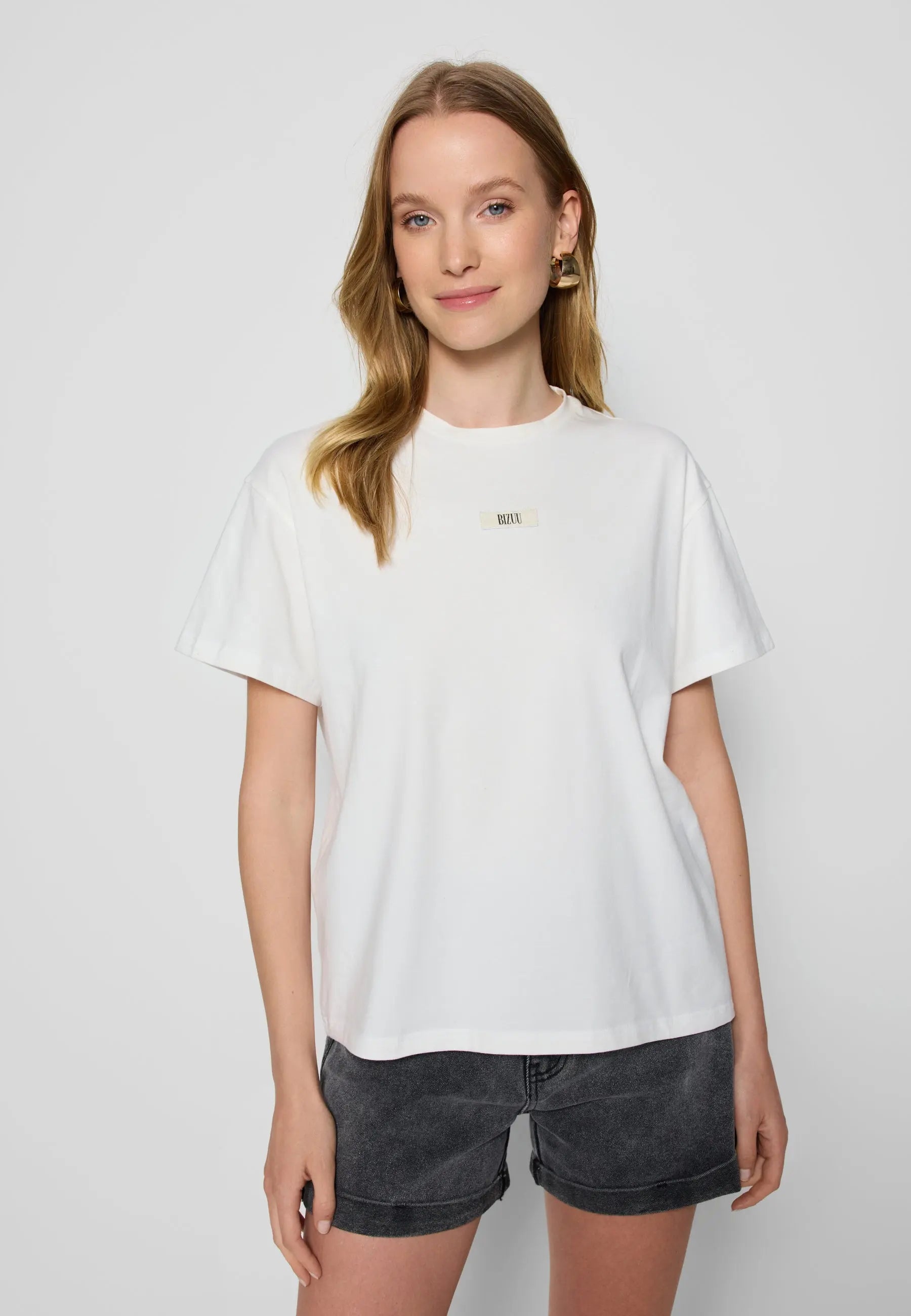 Modelka na białym tle w kremowym T-shircie TISHALLE i jeansowych szortach MENORCA.