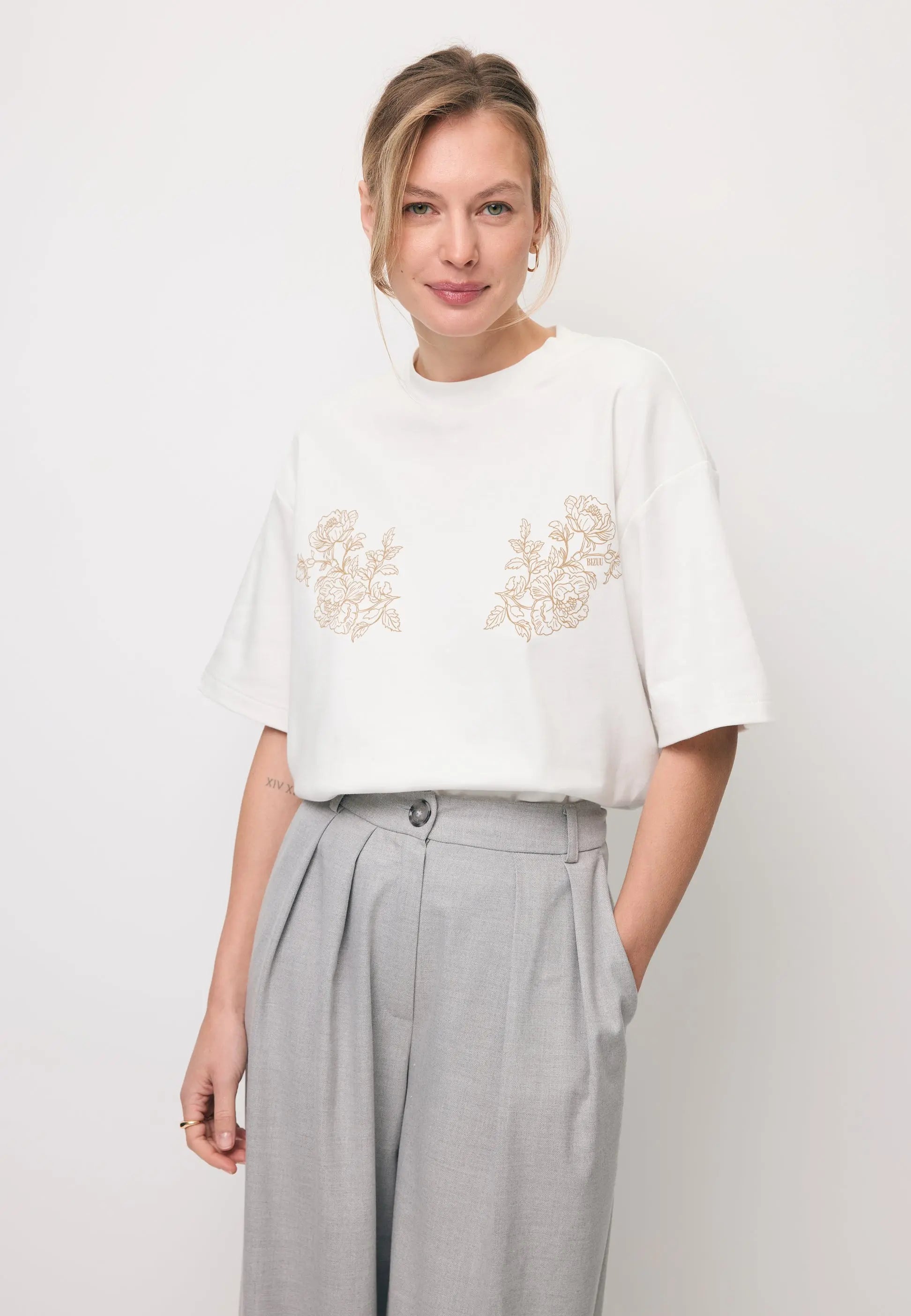 Modelka ubrana w kremową koszulkę oversize z ozdobnym nadrukiem FANYA oraz w szare spodnie BANOS.