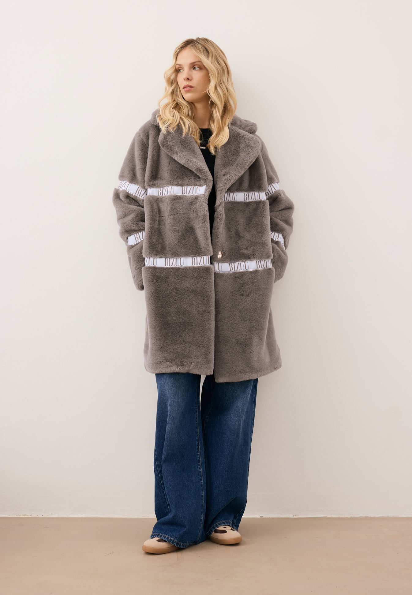 Modelka patrząca w bok, z rękoma w kieszeni futrzanego płaszcza FLORENCE z logo w kolorze taupe.