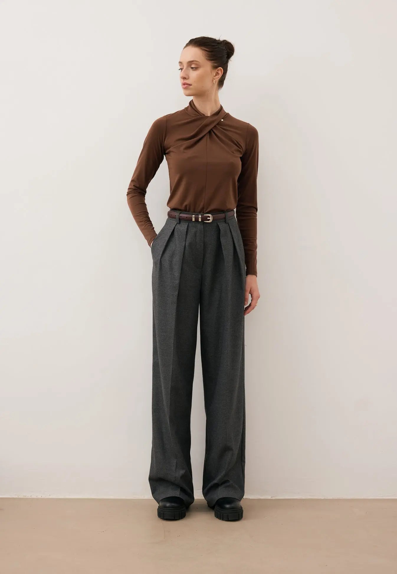 Modelka z jedną ręką w kieszeni spodni BANOS spogląda w bok. Ubrana w brązowy longsleeve PINOWA z zabudowanym dekoltem.